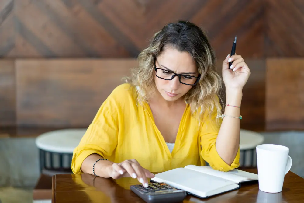Mujer calculando comisiones y gastos con una calculadora junto al portátil